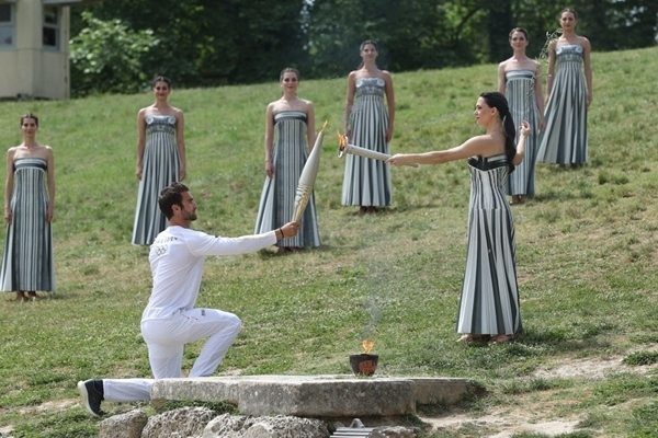 扮演最高女祭司的演員瑪麗·米娜（前右）點燃第一棒火炬手——希臘運動員、東京奧運會男子賽艇項目金牌得主斯特凡諾斯·恩圖斯科斯手中的火炬。新華社記者 趙丁喆 攝