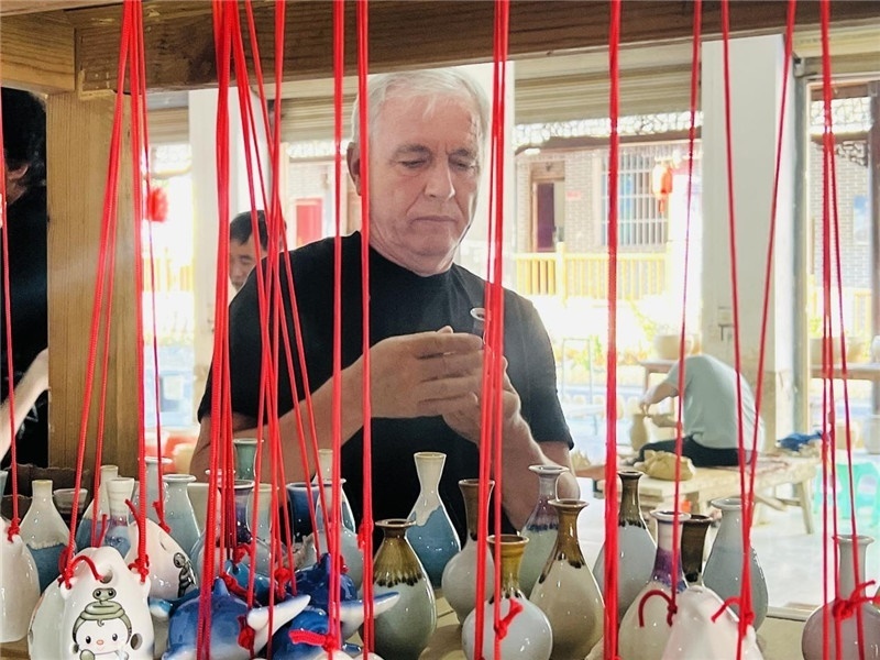 鄉(xiāng)愁如同一根根紅線，將游子與故鄉(xiāng)永遠(yuǎn)地牽連到一起?；逄账囄幕瘎?chuàng)意一條街的母氏陶藝館內(nèi)，紅線吊著陶瓶，思緒游走萬(wàn)里。摩爾多瓦“信息標(biāo)簽”通訊社社長(zhǎng)亞歷山大·塔納斯端詳著館內(nèi)展示的陶藝作品，并挑選出心儀的小擺件帶回摩爾多瓦，也帶走花茂村的“鄉(xiāng)愁”。（張亞欣攝）